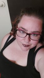 A femme presenting person with dark hair wearing a black dress and dark framed glasses looks up into the camera above them