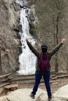 Elle stands on two large rocks with arms and legs outstretched, facing a distant waterfall with his back to the camera. His dark brown hair is in a braid. He is wearing a green jacket, a purple backpack, blue jeans, and hiking boots.