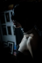 A femme body in side profile pictured in shadows, wearing a leather face mask and textured black corset top. Around her neck is a classic silver collar.
