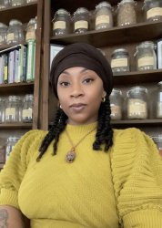 A  woman wearing a dark head wrap and yellow shirt sits in front of wooden shelves filled with glass jars of herbs and botanical materials. She has twists and is wearing a pendant necklace. Behind her are multiple rows of labeled glass filled jars and herb books.