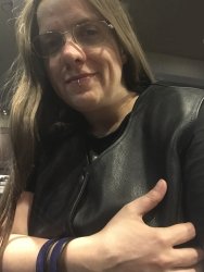 Femme presenting non-binary person with long hair, looking towards the camera, bracing themself, wearing glasses, a black leather vest, and a purple leather arm band.