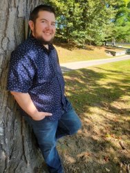 AJ has short dark hair and a beard leans against a large tree trunk in a sunny park, smiling toward the camera.  He wears a navy blue short-sleeved button-down shirt with a small white polka-dot pattern and blue jeans, with one hand in  his pocket and one foot propped against the tree. Behind him, a paved path curves past picnic tables and a grassy hillside  dappled with sunlight and shade from surrounding trees.
