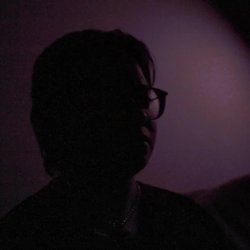 A pale, short-haired butch looks to the side of the camera. There is a dark purple background and he is in shadow.