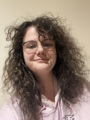The picture shows a smiling white non-binary person with long brown curly hair, glasses, septum and ear piercings. An o-ring day-collar around their neck. Bright pink jumper.