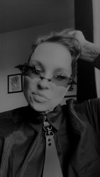 A black and white image of an androgynous individual in sunglasses and a a shirt and tie, featuring the face, neck, and shoulders only.