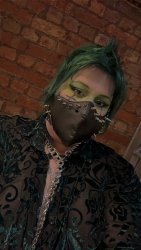 A photo of a white alternative person with a dark green fohawk, green and black eye makeup and a black leather look mask with silver spikes. They are wearing a dark green velvet patterned longsleeve mesh button up, that is mostly unbuttoned, with a long silver chain/leash poking through.