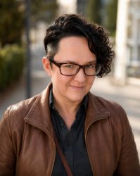 A person wearing tortoiseshell glasses and a brown leather jacket stands outdoors on a softly blurred sidewalk. They have short dark hair with closely cropped sides and longer curly hair swept to one side. A small septum piercing is visible. They are looking directly at the camera with a subtle, confident smile. The portrait is lit by warm natural light, creating soft highlights on the leather jacket and gentle shadows on the face. The background is out of focus, showing trees, greenery, and light-colored buildings, giving the image a professional environmental-portrait feel.