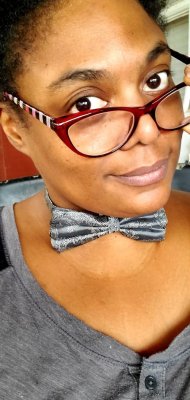 A woman with short natural hair wearing dark red cat-eye glasses, a gray paisley bow tie, and a gray henley shirt, smiling slightly at the camera.