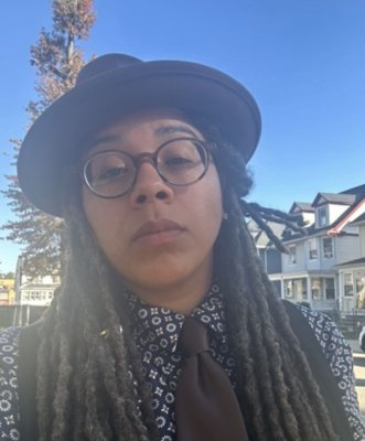 There is a Black person with a brown hat, brown round glasses, a brown tie, with a blue patterened shirt.
