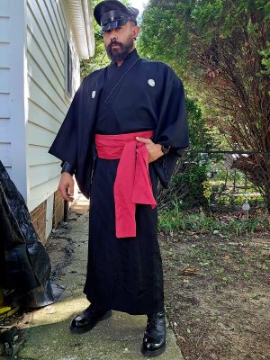 A man wearing a muir cap and a dark Kimono with a a red Obi. He is wearing Black boots while standing outside surrounded by trees.