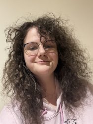 The picture shows a smiling white non-binary person with long brown curly hair, glasses, septum and ear piercings. An o-ring day-collar around their neck. Bright pink jumper.