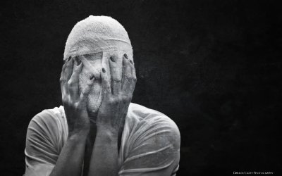 Black and white photograph of a person wrapped head to toe in white gauze bandaging, hands raised to cover their face, against a dark background. Photo by Darker Light Photography.