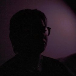 Photo of a white human in shadow, looking into the distance. He is wearing a collar and the background is a shadowy purple.