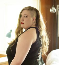 A curvy white woman in a black tank top looks over her left shoulder into the camera with red lips and a serious expression. Her light brown hair is shaved on the left side with the rest hanging down to the small of her back.