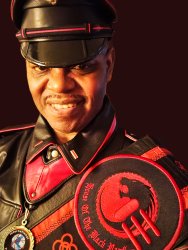 Sir Guy, a Black Leatherman in black leather cover with red trim, black leather uniform with red trim and black leather sash with House crest and red and black African Sankofa symbols