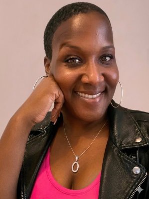 A smiling Black woman with closely cropped hair poses for a selfie, resting her cheek on one hand. She wears large hoop earrings, a silver pendant necklace, a bright fuchsia top, and a black leather jacket against a neutral background.