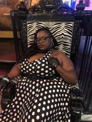 a leather gloved Black femme with locs in a vintage halter top black and white dress posed and seated in a zebra stripped Throne