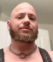 Close-up selfie of  a trans guy buzz cut, and with a short, well-groomed beard wearing a white, plush bunny hood with long, floppy ears that have pink inner lining. The setting is indoors with soft natural light coming from a window in the background.