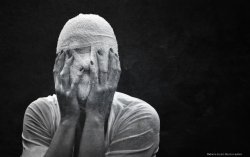 Black and white photograph of a person wrapped head to toe in white gauze bandaging, hands raised to cover their face, against a dark background. Photo by Darker Light Photography.