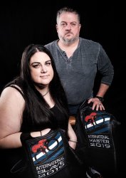 A man with grey hair and facial hair sits in a chair, while a woman with long dark hair kneels next to him They are holding leather vests with patches that says International Master and International slave. The man is wearing a grey long sleeve shirt with the sleeves pulled up and the woman is wearing a black sleeveless dress.