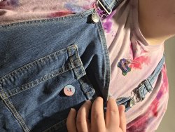 Nonbinary fat wearing tie dyed shirt and overalls displaying rainbow dog tag; "Trinket."