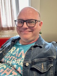 White, nonbinary bald person with black glass, a t-shirt, and jean jacket. T-shirt reads "Trans rights are human rights."