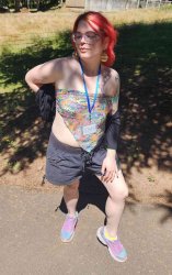 a white woman with bright red hair and glasses, wearing rainbow earrings, a colorful 'my little pony' handkerchief tube top, black shorts, and pink & blue sneakers. she is outdoors on a sunny day, standing in the shade of a tree, glancing down and to the side with a smirk, posing with a hand on her hip.