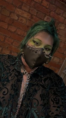 A photo of a white alternative person with a dark green fohawk, green and black eye makeup and a black leather look mask with silver spikes. They are wearing a dark green velvet patterned longsleeve mesh button up, that is mostly unbuttoned, with a long silver chain/leash poking through.