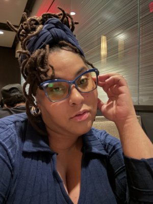 Close-up selfie of a woman with locs wrapped in a navy headband, wearing blue-framed glasses and a navy top while seated indoors.