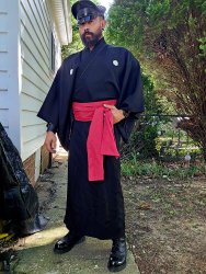 A man wearing a muir cap and a dark Kimono with a a red Obi. He is wearing Black boots while standing outside surrounded by trees.