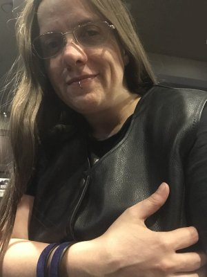 Femme presenting non-binary person with long hair, looking towards the camera, bracing themself, wearing glasses, a black leather vest, and a purple leather arm band.