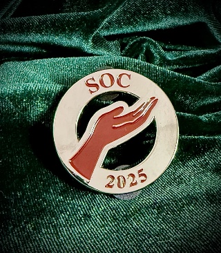 A round silver pin against a green velvet backdrop. The pin has the letters S O C across the top, 2025 across the bottom, and a hand held out in offering in the middle, all in red enamel. There are two see-through cutouts above and below the main part of the hand, making it look like it floats in space.