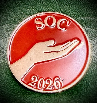 A round silver pin against a green velvet backdrop. The pin has the letters S O C across the top, 2026 across the bottom, and a hand held out in offering in the middle, all in red enamel.